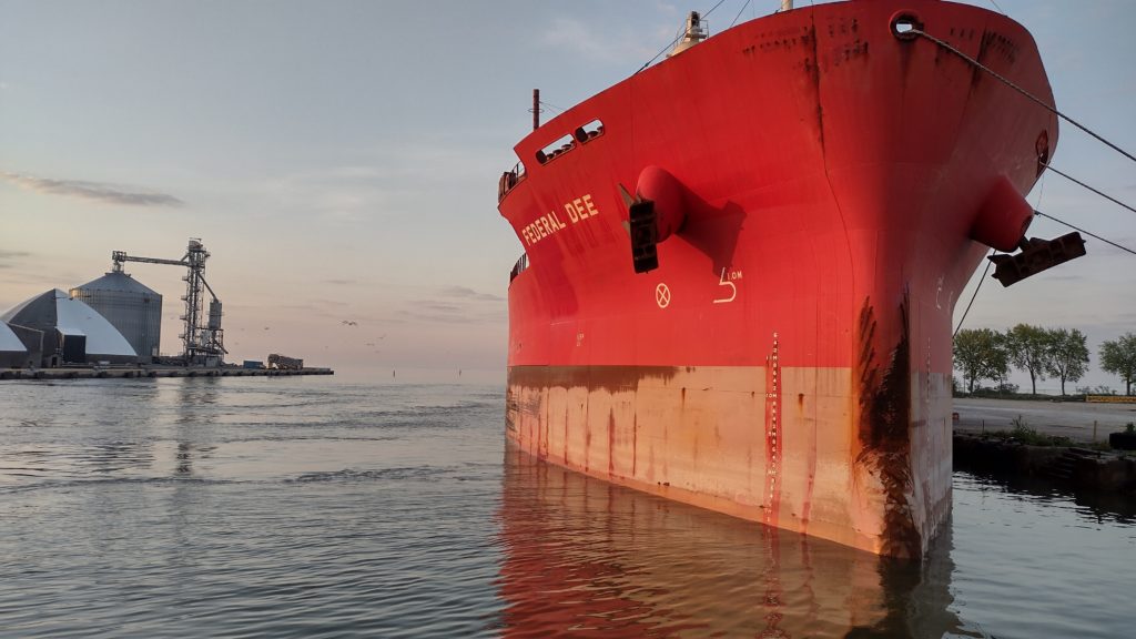 The bow of a large red ship faces the camera, with the faint glow of sunrise behind it