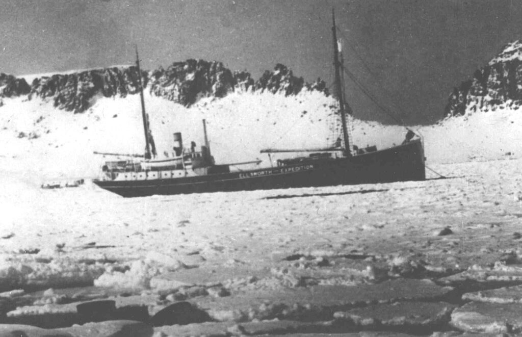 a black and white photo of a ship, near Antarctica with Mountains behind it. The ship is traversing from left to right.