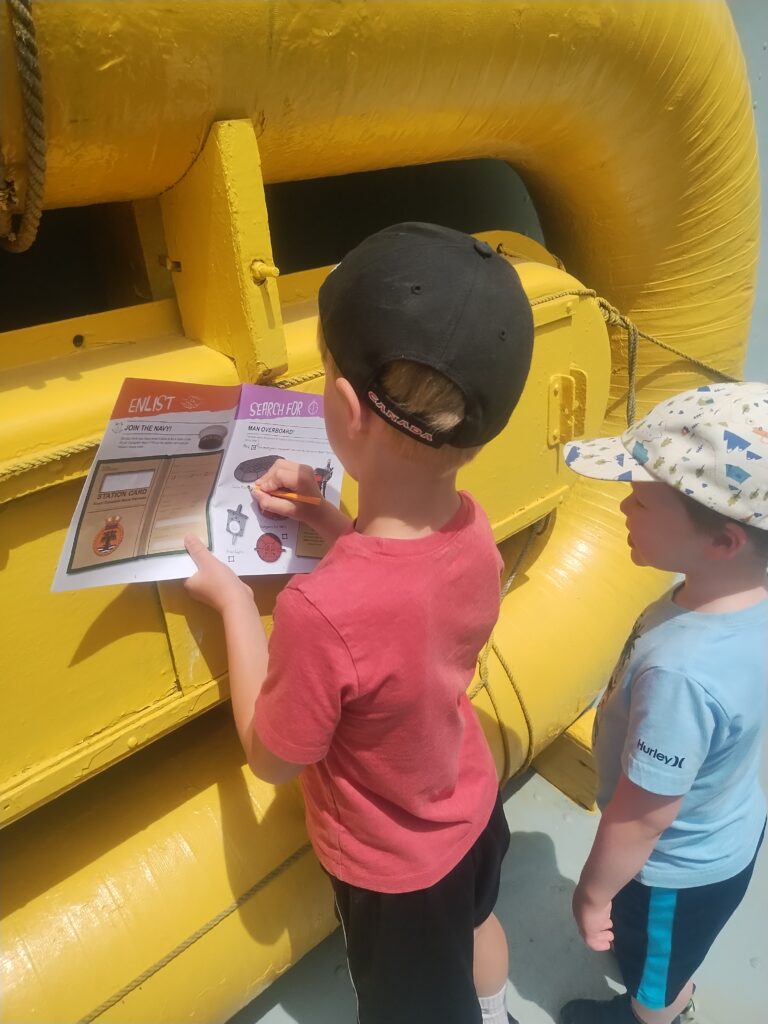 two boys look at a bright yellow Carley Float. The older one checks off the find in the activity book