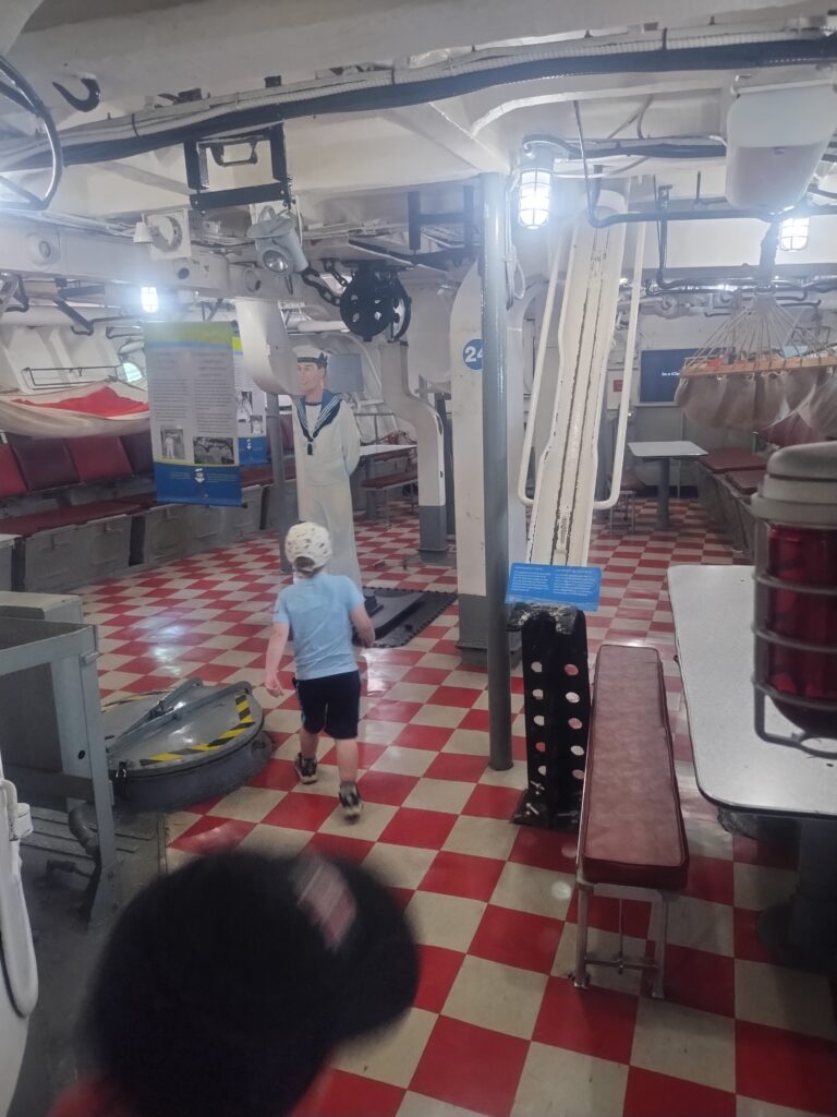 A small boy enters a space that contains tables, hammocks, and the ammunition hoists for the forward 4" guns