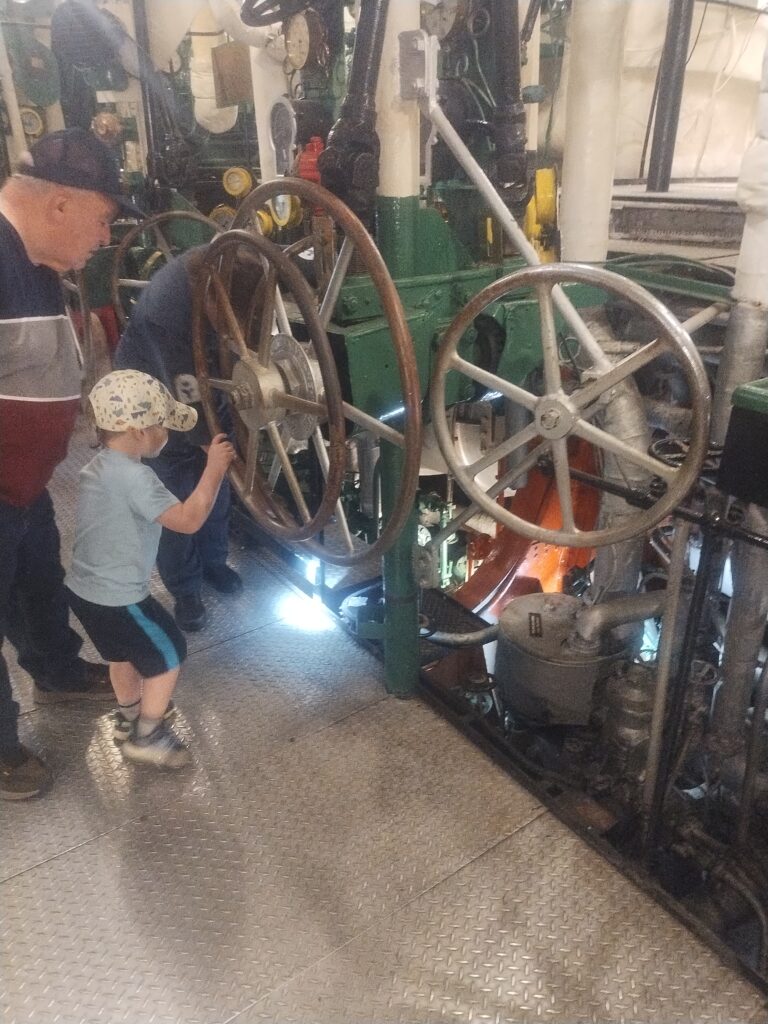 A woman in coveralls explaining steam engine functions to a small boy and an older man