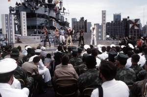 a group of sailors in the foreground watch the musical act' Village People' perform